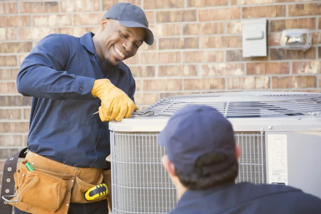 A cheerful male HVAC technician, wearing a dark blue shirt, a cap, a leather tool belt, and yellow work gloves, is smiling while using a wrench to work on a large outdoor air conditioning condenser unit. He is interacting with a second technician whose back is to the camera.